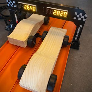 Könnte beinhalten: Zwei Holz-Rennwagen auf einer orangefarbenen Rennstrecke, positioniert unter einer schwarzen Digitaluhr, die die Rennzeiten anzeigt. Die Wagen sind helles Holz mit schwarzen Rädern. Die Uhr zeigt "2.640" und "2.820" an. Das PlayRobotics-Logo ist sichtbar.