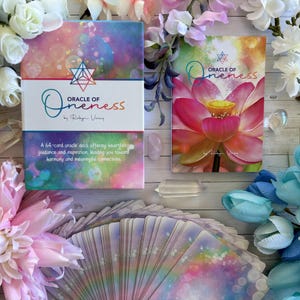 May include: A 64-card oracle deck called "Oracle of Oneness" by Robyn Voiers. The deck features a colorful, abstract design with a pink lotus flower on the guidebook cover. The cards are spread out on a white wooden surface with crystals and flowers.