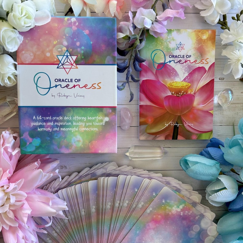 May include: A 64-card oracle deck called "Oracle of Oneness" by Robyn Voiers. The deck features a colorful, abstract design with a pink lotus flower on the guidebook cover. The cards are spread out on a white wooden surface with crystals and flowers.