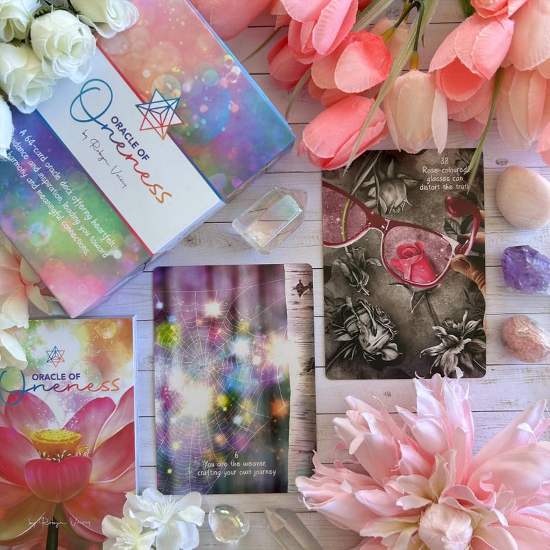 May include: A set of oracle cards with a pink and purple color scheme. The cards feature images of a spider web, a rose, and a lotus flower. The cards are titled "Oracle of Oneness" and are by Robyn Vovrey.