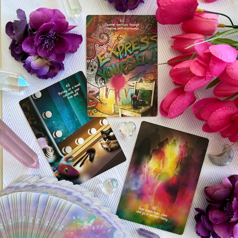 May include: Three oracle cards with colorful designs and text. The first card has a graffiti-style design with the text "Express Yourself". The second card has a blue background with a mask and makeup brushes. The third card has a colorful, abstract design with the text "Soul connections come and go in perfect timing".