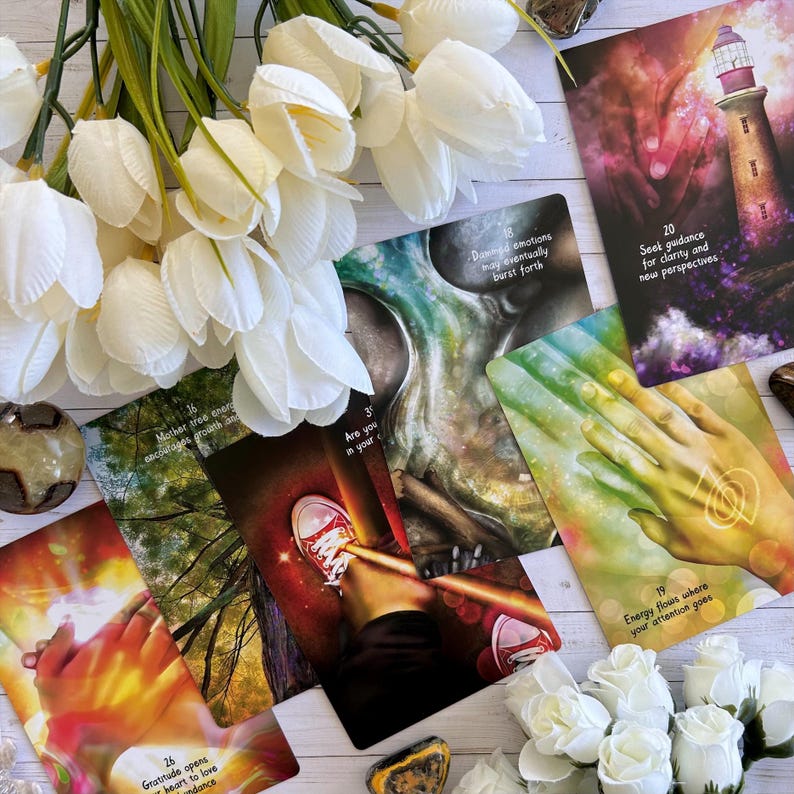 May include: A set of colorful oracle cards with images of hands, trees, and a lighthouse. The cards have numbers and text on them, including "Mother tree energy encourages growth and", "Damned emotions may eventually burst forth", "Seek guidance for clarity and new perspectives", "Are you in your", "Gratitude opens your heart to love and dance", "Energy flows where your attention goes".