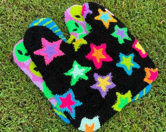 May include: A tufted rug featuring a black background with a pattern of multi-colored stars in shades of neon pink, yellow, blue, purple, and green. The rug has a rounded, organic shape with a secondary layer showing a green alien face design.