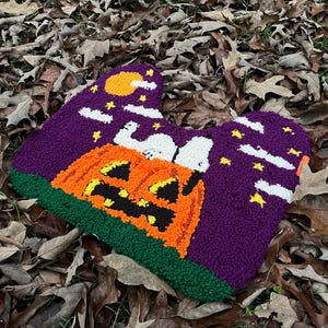 May include: A Halloween-themed rug in a heart shape, featuring a jack-o'-lantern, Snoopy, and Woodstock. The rug has a purple background with stars and a moon, and a green border. The design is made with textured yarn.