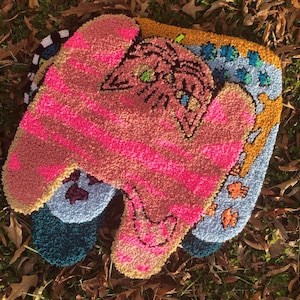 May include: A colorful rug made of looped yarn with a pink, blue, and yellow design. The rug features a pink and white striped design with a pink and white cat face in the center.