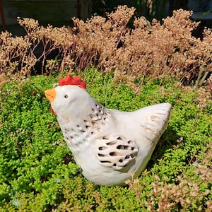 Könnte beinhalten: Eine weiße Keramik-Hühnerfigur mit rotem Kamm und orangefarbenem Schnabel. Das Huhn hat eine strukturierte Oberfläche mit ausgeschnittenen Details. Es steht inmitten von grünem Laub und braunen Trockenpflanzen.