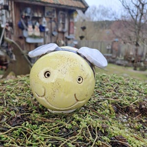 Könnte beinhalten: Keramik-Bienenfigur mit lächelndem Gesicht, gelbem Körper und weißen Flügeln. Die Biene hat schwarze Streifen und Augendetails. Die Figur steht auf grünem Moos.