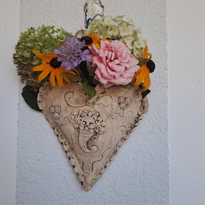 Könnte beinhalten: Ein herzförmiger Wandübertopf aus beigefarbener, strukturierter Keramik mit geprägten Blumen- und Rankenmustern. Der Übertopf ist mit einem bunten Arrangement aus grünen Hortensien, gelben Sonnenhüten, einer violetten Skabiose und einer rosa Rose gefüllt.