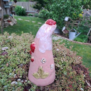Könnte beinhalten: Ein rosa Keramik-Gartenzwerg mit Erdbeermotiv. Der Zwerg hat eine gebogene Mütze mit einer strukturierten roten Erdbeere oben und mehrere kleinere Erdbeeren und Blumen am Körper. Am Boden befindet sich ein grünes Blatt.