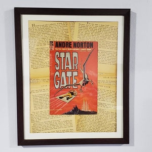 May include: A framed vintage book cover featuring a red, white, and black illustration of a flying creature with a man in a spacesuit falling from the sky. The book title is "Star Gate" and the author is Andre Norton. The text "Norton again demonstrates a superb talent" is also visible.