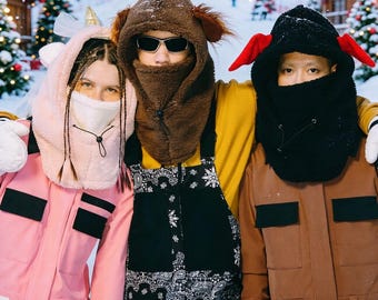 Könnte beinhalten: Drei Fleece-Sturmhauben mit Tiermotiven: eine hellrosa Einhorn-Kapuze mit goldenem Horn, eine braune Bären-Kapuze mit Ohren und eine schwarze Kapuze mit roten Ohren. Jede Kapuze verfügt über eine integrierte Gesichtsmaske mit Kordelzug.