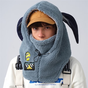 Puede incluir: Un calentador de cuello azul de forro polar con capucha y orejeras. El calentador de cuello tiene un parche amarillo y negro con el texto "OH! ANON" y un parche blanco y negro con una cara de dibujos animados. El texto "Try A Little Harder To be A Little Better." está impreso en la parte inferior del calentador de cuello.