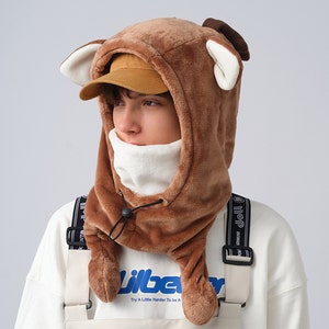 May include: A brown fleece hooded hat with deer ears. The hat has a white fleece lining and a black drawstring. The hat is worn by a person wearing a white shirt and a black strap.