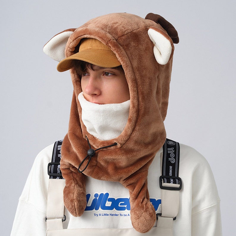 May include: A brown fleece hooded hat with deer ears. The hat has a white fleece lining and a black drawstring. The hat is worn by a person wearing a white shirt and a black strap.