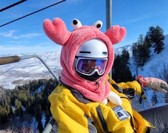 Könnte beinhalten: Ein leuchtend rosa Plüsch-Helmüberzug in Krabbenform mit zwei großen Scheren, zwei hervorstehenden Augen und einem Kordelzug. Der Überzug wird über einem weißen Skihelm mit verspiegelter Skibrille getragen, dazu eine gelbe Jacke und beige Handschuhe.