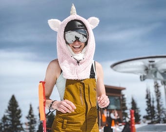 Könnte beinhalten: Eine hellrosa Einhorn-Kapuze aus Plüsch mit Ohren und Horn, getragen über einem schwarzen Helm und verspiegelter Skibrille. Das Outfit besteht aus einer senfgelben Latzhose mit Frontreißverschluss über einem hellen Tanktop.