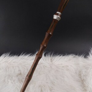 May include: A handcrafted wooden wand with a dark brown finish and silver accents. The wand features a decorative orb at the top and green painted details. The wand is displayed against a white furry background.