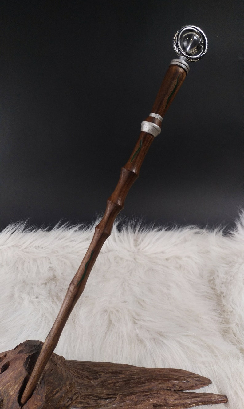 May include: A handcrafted wooden wand with a dark brown finish and silver accents. The wand features a decorative orb at the top and green painted details. The wand is displayed against a white furry background.