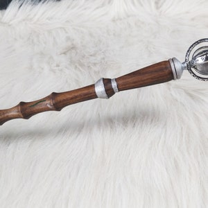 May include: A handcrafted wooden wand with a dark brown finish, accented with silver bands and a decorative sphere at the top. The wand has a textured, bamboo-like design with green accents.