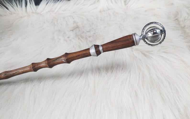 May include: A handcrafted wooden wand with a dark brown finish, accented with silver bands and a decorative sphere at the top. The wand has a textured, bamboo-like design with green accents.