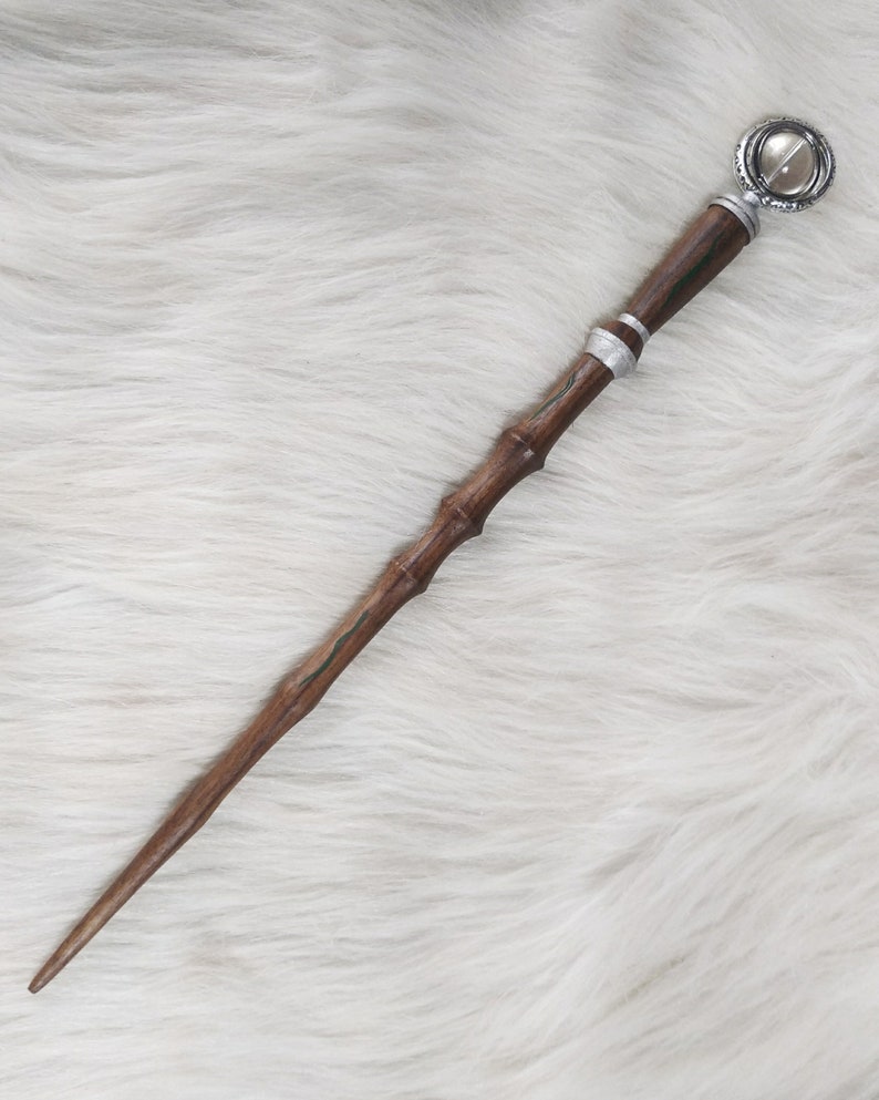 May include: A wooden wand with a dark brown finish, featuring a silver band and a decorative, circular top with a clear stone. The wand has a textured, bamboo-like design and green accents. The wand is approximately 14 inches long.