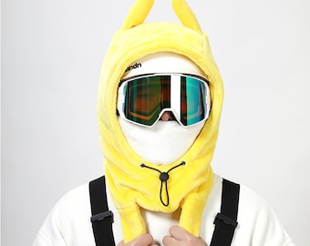Könnte beinhalten: Eine gelbe Skimaske mit Hasenohren, einer weißen Skibrille und einer weißen Gesichtsbedeckung. Die Maske hat einen Kordelzug und wird über einem weißen Helm getragen. Schwarze Hosenträger sind sichtbar.