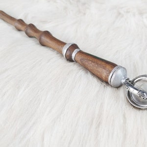 May include: A wooden wand with a dark brown finish, silver accents, and a decorative orb at the end. The wand has a detailed, carved design and is set against a white, fluffy background.
