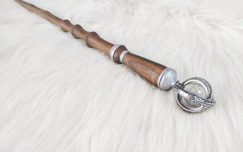 May include: A wooden wand with a dark brown finish, silver accents, and a decorative orb at the end. The wand has a detailed, carved design and is set against a white, fluffy background.