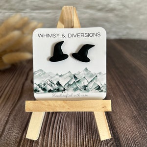 May include: A pair of black witch hat stud earrings on a white card with the text "Whimsy & Diversions" and "Handcrafted with care".