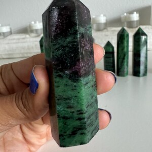 May include: A polished zoisite crystal tower with a green and black pattern, held in a hand. The crystal has a dark purple hue at the top. Several other smaller crystals are visible in the background.