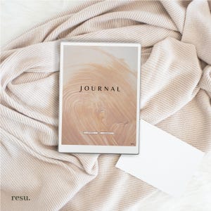 May include: A white journal with a brown and beige abstract design on the cover. The text "JOURNAL" is printed in black on the cover. The journal is lying on a white and beige knitted blanket.