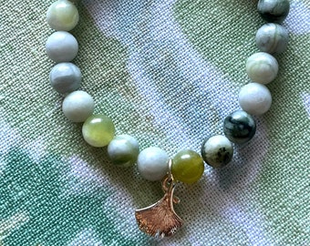 May include: A green and white beaded bracelet with a silver ginkgo leaf charm. The bracelet is made of natural stones and is a beautiful and unique piece of jewelry.