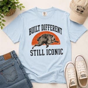 May include: A light blue t-shirt featuring a graphic of a running raccoon in front of an orange semi-circle. The text "BUILT DIFFERENT" is arched above the raccoon, and "STILL ICONIC" is printed below in a bold, dark, sans-serif font.