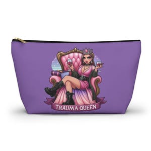 Peut inclure: Une pochette zippée violette avec une illustration d'une femme en robe rose et couronne, assise sur un trône, tenant des verres à vin. Le texte "TRAUMA QUEEN" est sur une bannière. La pochette a une fermeture éclair dorée.