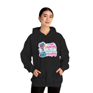 Op de afbeelding: Zwarte hoodie met een kleurrijke afbeelding van een lachende beker en de tekst "I'M NOT CHRONICALLY ILL; I'M MEDICALLY FASCINATING". Het ontwerp maakt gebruik van heldere kleuren en speelse lettertypen, perfect voor wetenschapsliefhebbers.