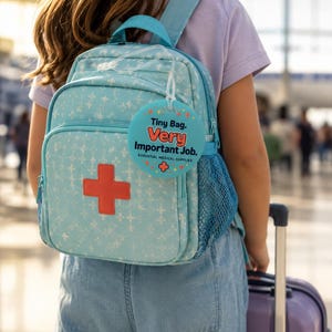 May include: A light blue backpack with a repeating white cross pattern and a large red cross patch on the front pocket. A round teal tag is attached, featuring the text: Tiny Bag. Very Important Job. Essential Medical Supplies, with a small red cross.