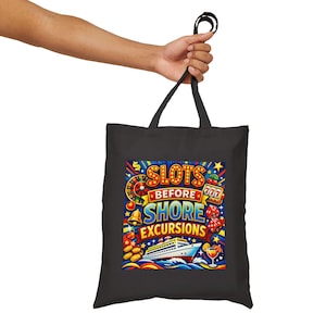 May include: Black tote bag with a colorful graphic that reads "SLOTS BEFORE SHORE EXCURSIONS." The design includes a roulette wheel, dice, a cruise ship, and other casino-themed elements. The bag is held by a person.
