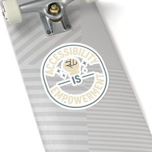 May include: A round sticker with the words "ACCESSIBILITY IS EMPOWERMENT" in a circular design. The sticker features a fist graphic and starbursts in a cream and teal color scheme, set against a gray and white striped background.