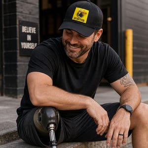 May include: Black baseball cap with a yellow square patch featuring text. The cap is worn by a person wearing a black t-shirt and shorts. The person is sitting on concrete steps. A prosthetic leg is visible.