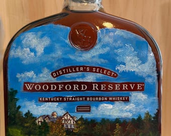 May include: A Woodford Reserve Kentucky Straight Bourbon Whiskey bottle featuring a hand-painted landscape scene over the label. The artwork depicts a green golf course, trees, and a building under a blue sky with white clouds. Text includes Distiller's Select.