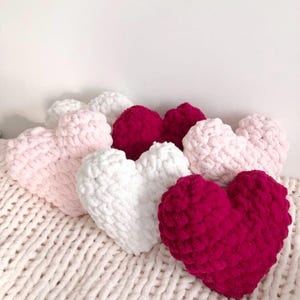 May include: Five heart-shaped knitted pillows in pink, white, and red. The pillows are arranged in a row on a white knitted blanket.