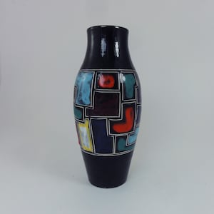 Op de afbeelding: Een hoge, donkerblauwe keramische vaas met een glanzende afwerking. De vaas heeft een geometrisch patroon van kleurrijke vormen in rood, blauw, geel en groen, omlijnd in wit. De vaas heeft een smalle hals en een bredere basis.
