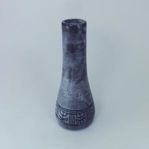 May include: A tall, slender vase with a dark grey and light grey speckled finish. The vase has a flared base and a narrow neck. A decorative band with a repeating floral pattern encircles the base. The vase is approximately 20 cm tall.