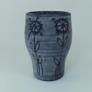 May include: A grey ceramic tumbler with a floral design. The tumbler features two black flower illustrations with stems and leaves. The ceramic has a textured appearance and a slightly tapered shape.