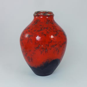 Op de afbeelding: Een keramische vaas met een afgeronde body en een smalle hals. De vaas is voornamelijk rood met een gespikkeld patroon en een donkerblauwe basis. De rand heeft een zilverkleurige rand. Deze decoratieve vaas is geschikt voor woondecoratie.
