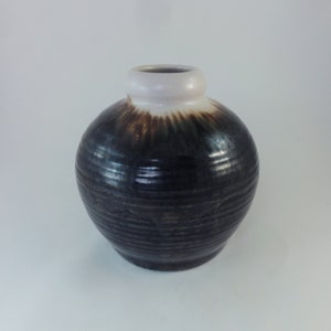 May include: A round, black ceramic vase with a white rim. The vase has a brown glaze and a textured surface.
