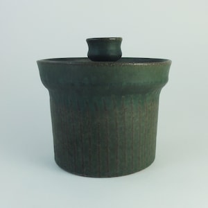 Op de afbeelding: Een groene keramische pot met een deksel. De pot heeft een getextureerd oppervlak en een licht afgeronde vorm. Het deksel is een apart onderdeel en heeft een eenvoudig, afgerond ontwerp.