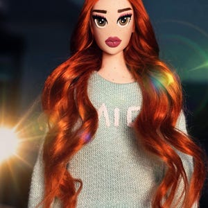 May include: A doll with long, wavy, vibrant red hair, freckles, and large brown eyes. The doll wears a light blue long-sleeved sweater with partial white text visible on the chest and a dark gray textured skirt. The doll has a neutral expression.