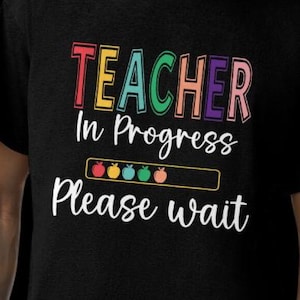 May include: Black t-shirt with the words "TEACHER In Progress Please wait" in colorful, playful lettering. The word "TEACHER" is in rainbow colors. Below the text is a loading bar with apple icons.