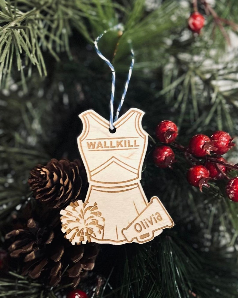 May include: A wooden Christmas ornament shaped like a cheerleader outfit with a pom-pom and megaphone. The ornament is personalized with the text "WALLKILL" and "Olivia".